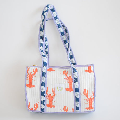 The Sara Fitz Purse, Lobster is a handmade rectangular cotton shoulder bag from India with blue handles and trim, quilted texture, and playful orange lobster and white seashell patterns on a white background.
