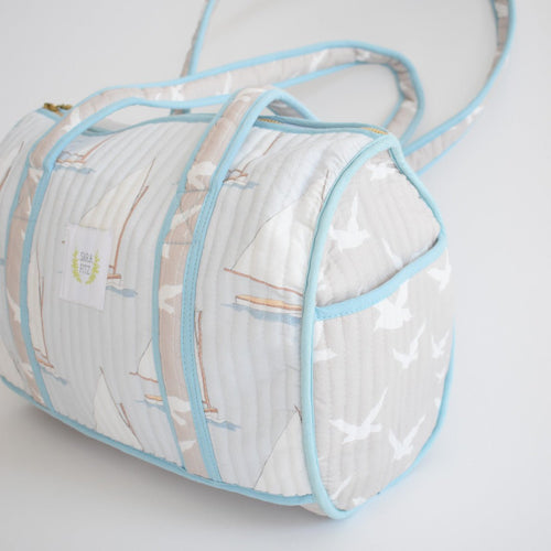 The Sara Fitz Purse, Sailboat is a handmade light blue and white quilted shoulder bag from India with sailboat and seagull patterns, blue piping, two handles, and a shoulder strap, shown on a white background.