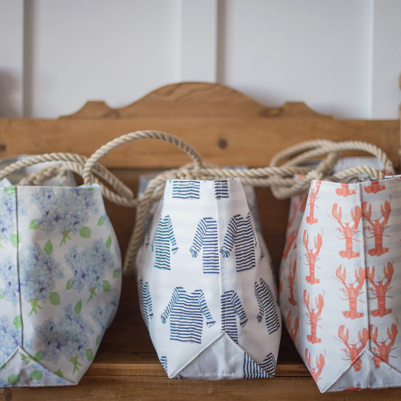 Three fabric bags with different prints—blue hydrangeas, navy striped shirts, and orange lobsters—sit on a wooden bench with a thick rope handle threaded through them.