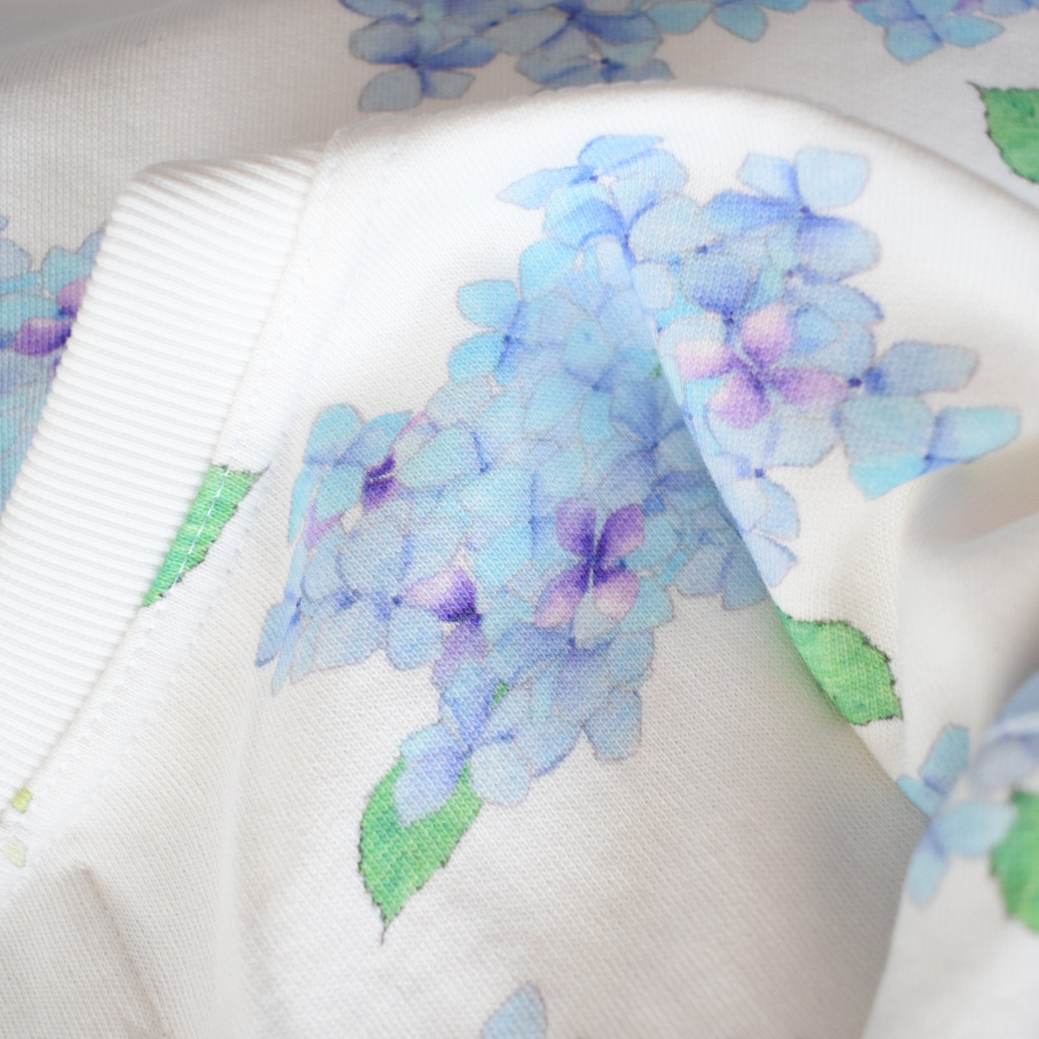 A close-up of the Hydrangea Sweatshirt by Sara Fitz shows soft, slightly wrinkled cotton featuring a floral print with clusters of blue and purple hydrangea flowers and green leaves, custom crafted in India.