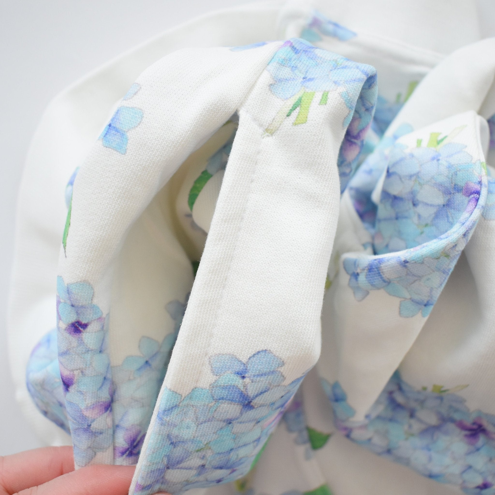 A close-up of the Sara Fitz Hydrangea Sweatshirt shows soft cotton fabric with light blue and purple hydrangea flowers, gently gathered and held by a hand in the corner.