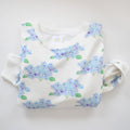 The Sara Fitz Hydrangea Sweatshirt, custom crafted in India from white cotton and featuring a light blue and purple hydrangea pattern with green leaves, is displayed on a white background.