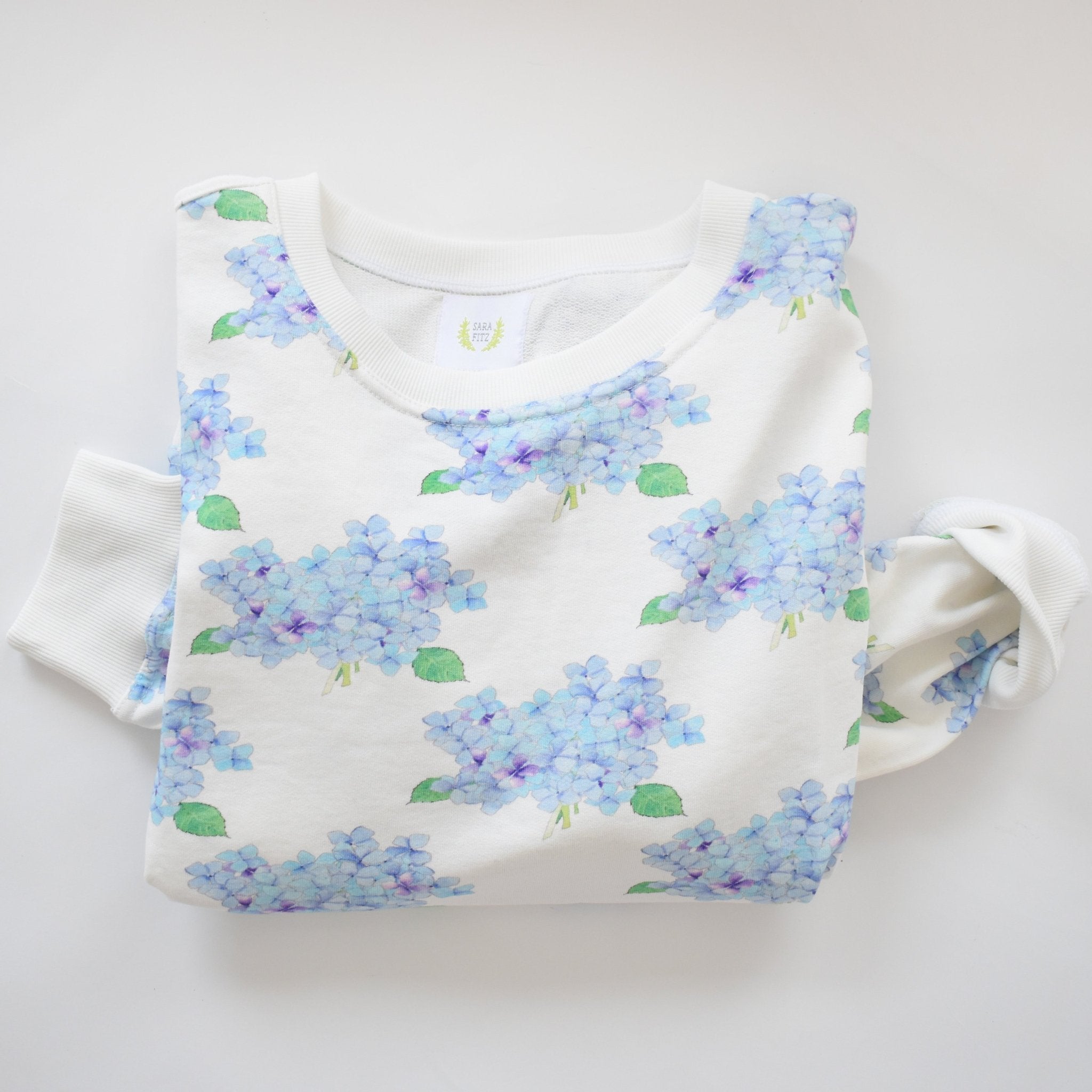 The Sara Fitz Hydrangea Sweatshirt, custom crafted in India from white cotton and featuring a light blue and purple hydrangea pattern with green leaves, is displayed on a white background.