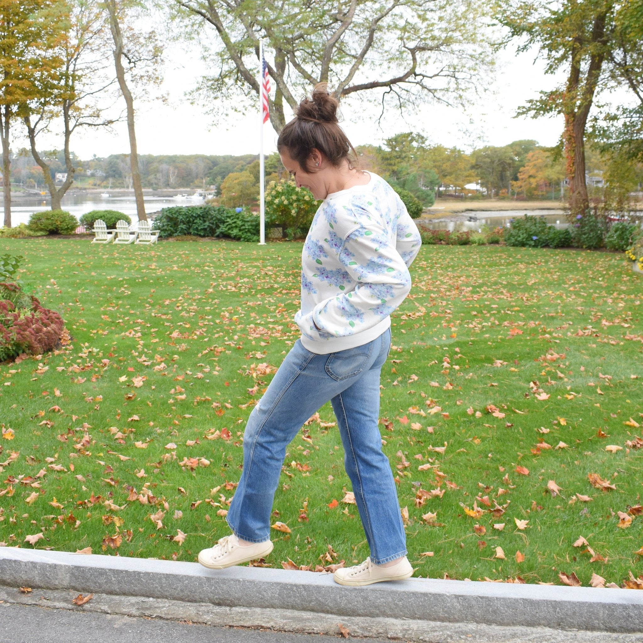 A person with a bun hairstyle wears the Sara Fitz Hydrangea Sweatshirt, blue jeans, and cream sneakers, standing on a curb in a green lawn with autumn leaves, trees, and a river in the background.