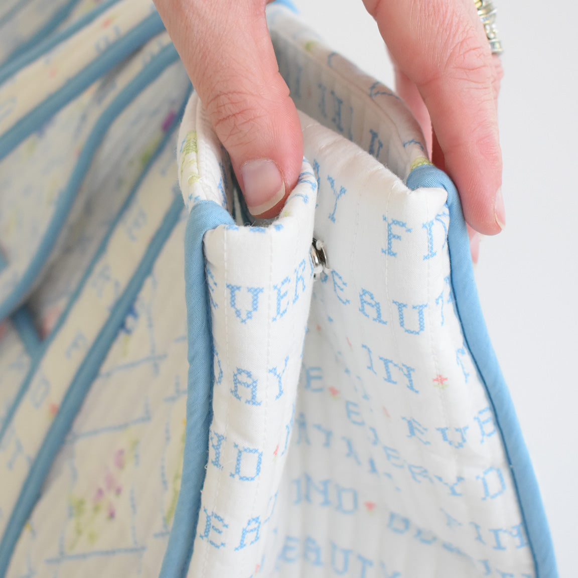 A hand with a silver ring holds the edge of the Sara Fitz Expandable Tote, Cross Stitch Floral, a quilted cotton bag with blue trim, stitched blue text, and a visible metal snap closure inside.