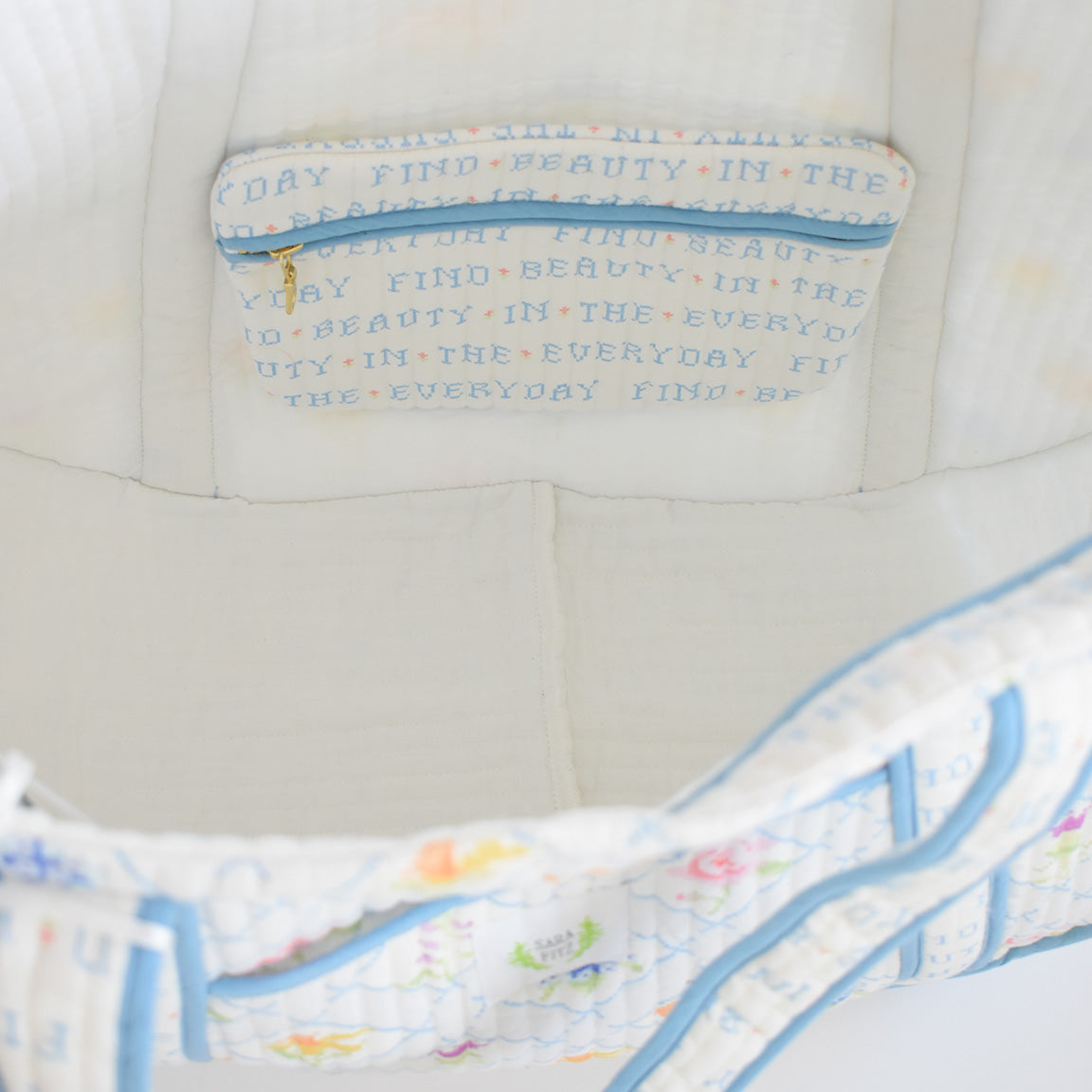 A close-up of the Sara Fitz Expandable Tote, Cross Stitch Floral shows its white quilted cotton interior with blue trim and an inner pocket embroidered with “FIND BEAUTY IN THE EVERYDAY” in colorful letters.