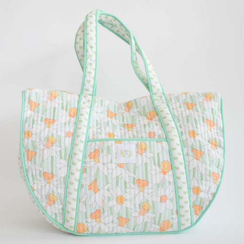 The Sara Fitz Expandable Tote, Daffy, is a large quilted cotton bag with light green stripes, white and orange daisies, green-trimmed edges, long handles, an exterior pocket, and a small white front label.