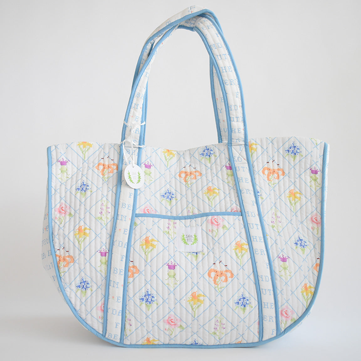 The Sara Fitz Expandable Tote, Cross Stitch Floral, features a quilted cotton design with pastel floral patterns, blue diamond lines on a white background, light blue edging, two handles, and a front pocket.