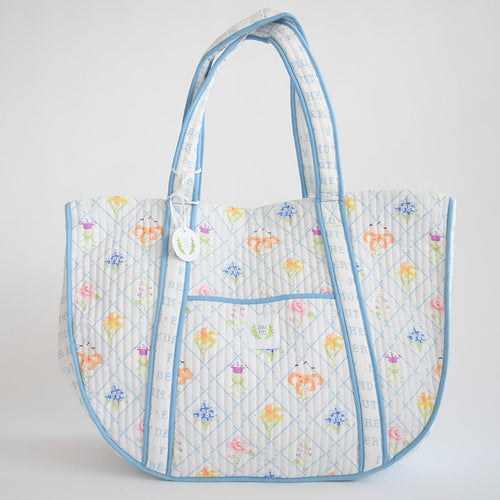 The Sara Fitz Expandable Tote, Cross Stitch Floral, features a quilted cotton design with pastel floral patterns, blue diamond lines on a white background, light blue edging, two handles, and a front pocket.