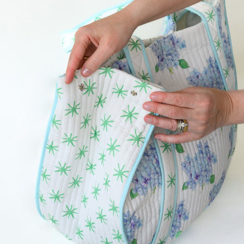 Two hands fasten a green star-patterned flap onto the Sara Fitz Expandable Tote, Hydrangea, which features blue and purple hydrangea designs, light blue trim, and a white background.