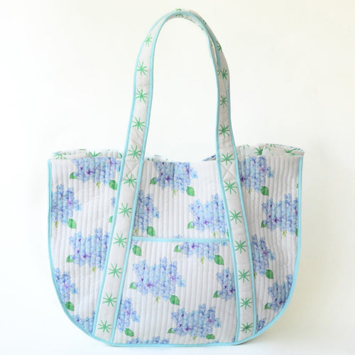 The Sara Fitz Expandable Tote, Hydrangea, features quilted cotton with a light background, blue and purple hydrangea florals, light green leaves, and aqua blue trim and straps with a star-like design.
