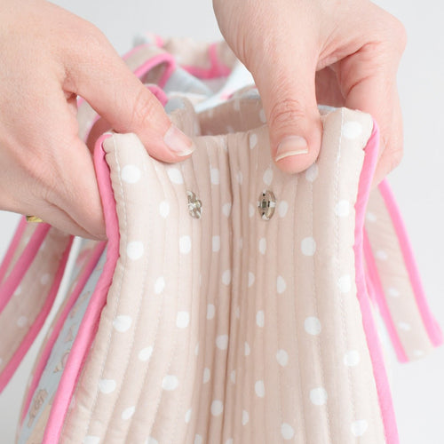 Close-up of two hands fastening metal snap buttons on the Sara Fitz Expandable Tote, Shell—a soft, quilted cotton bag with white polka dots and pink edging.
