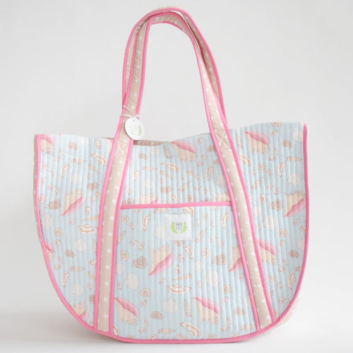 The Sara Fitz Expandable Tote, Shell, is a large quilted cotton bag with pink trim, two handles, a front pocket, and features pastel seashell and surfboard designs on light blue. A small white tag hangs from one handle.