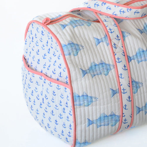 The Sara Fitz Weekender Tote, Blue Fish, is a quilted cotton bag with pink trim and blue fish and anchors on white. It features sturdy handles and a zippered closure—perfect for stylish weekends away.