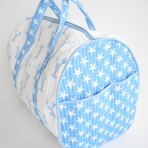 The Sara Fitz Weekender Tote, Blue Lobster, is a quilted cotton travel bag with a blue and white lobster print body and a white starfish pattern end pocket. It features two handles and a zipper closure.