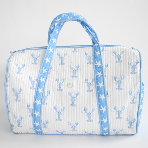 The Sara Fitz Weekender Tote, Blue Lobster, is a quilted white cotton bag with light blue handles and trim, adorned with blue lobsters and starfish. It features a zip closure and a small green-and-white label on the front.