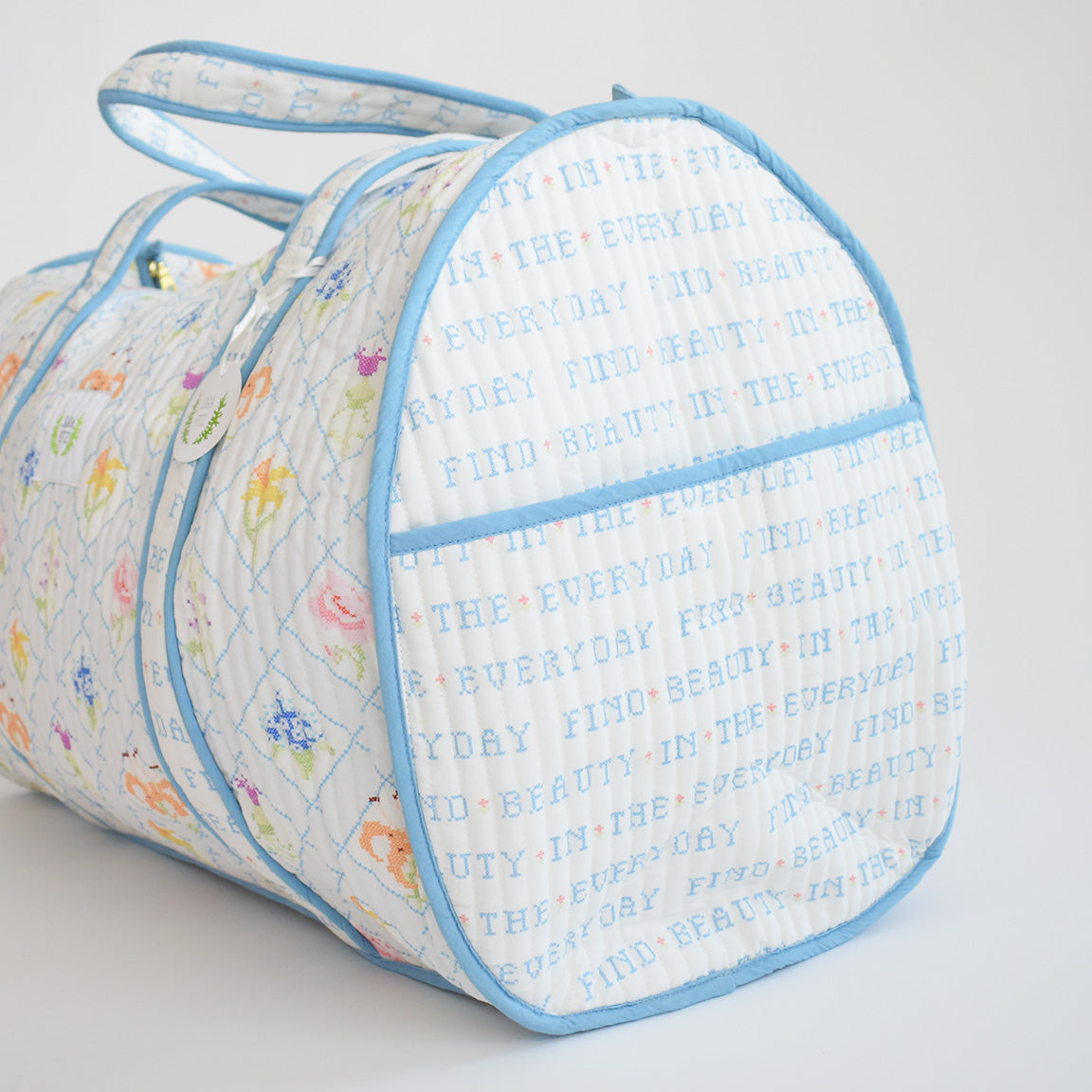 The Sara Fitz Weekender Tote, Cross Stitch Floral, is a white quilted travel bag with light blue trim and handles, featuring pastel floral designs and “FIND BEAUTY IN THE EVERYDAY” embroidered in blue letters.