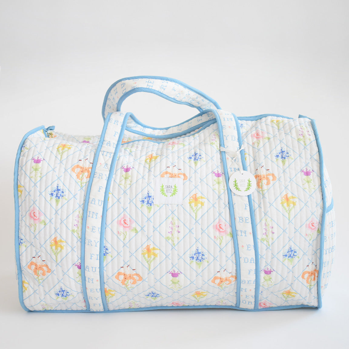 The Sara Fitz Weekender Tote, Cross Stitch Floral, is a quilted cotton travel bag with light blue trim and handles, featuring pastel floral and geometric patterns on white. A round white tag with green writing hangs from one handle.