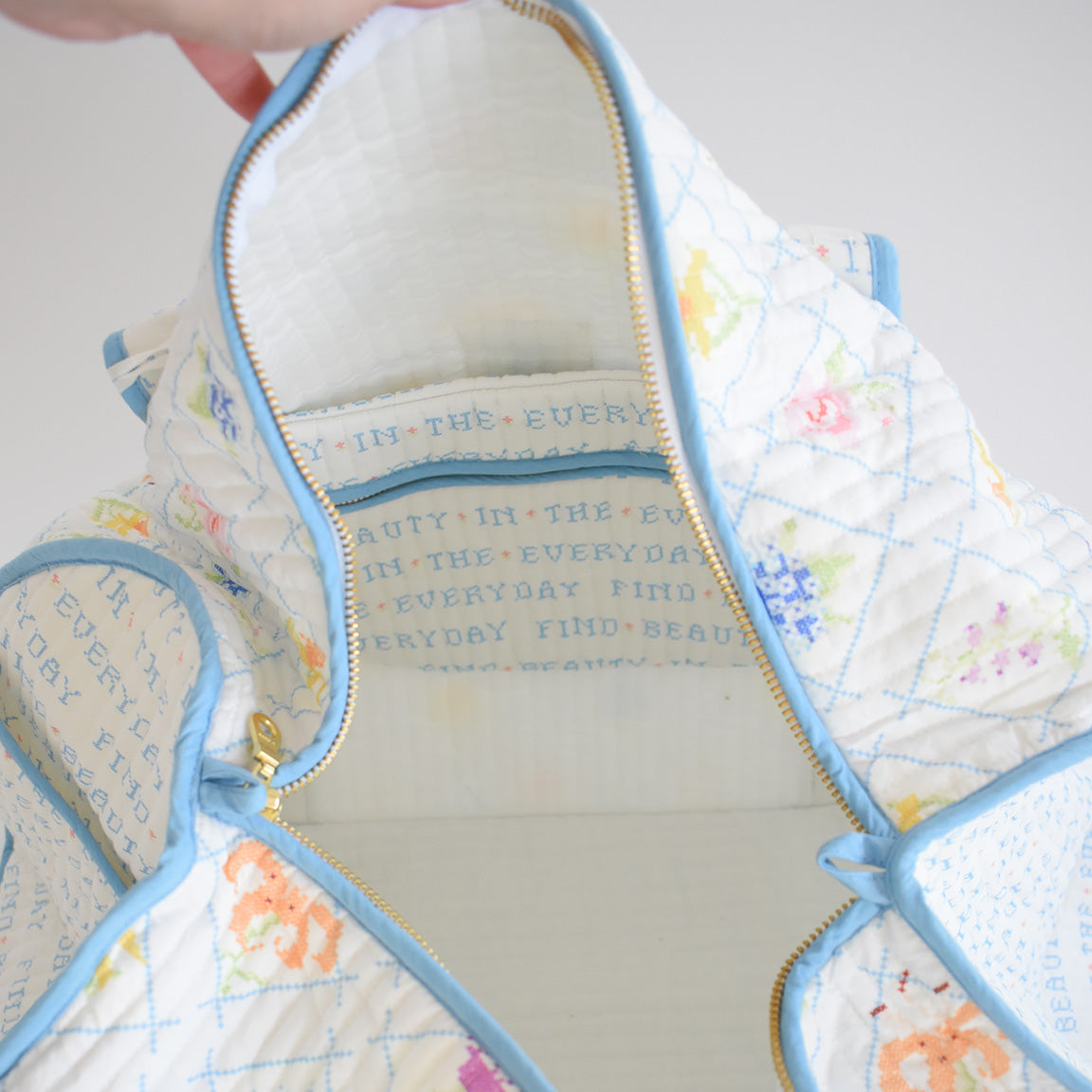 A close-up of the Sara Fitz Weekender Tote in Cross Stitch Floral shows its quilted white cotton, pastel floral design, and blue trim. The unzipped interior features repeating text: FIND BEAUTY IN THE EVERYDAY.