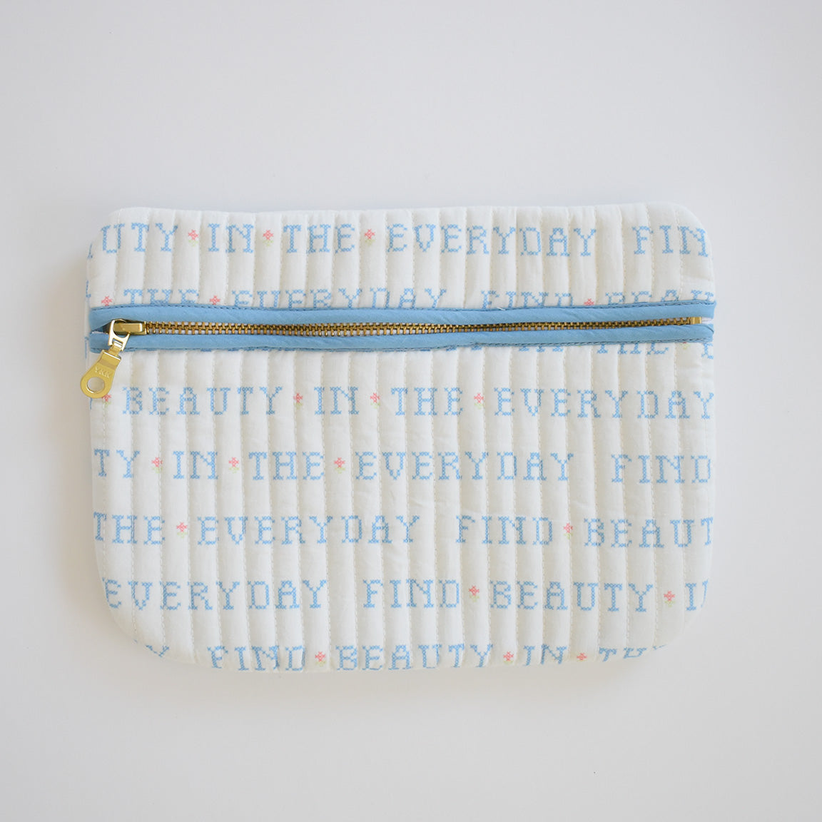 The Sara Fitz Weekender Tote, Cross Stitch Floral, is a white quilted cotton bag with a blue zipper and “FIND BEAUTY IN THE EVERYDAY” printed in light blue with pink cross stitch details—ideal for carrying your essentials on the go.