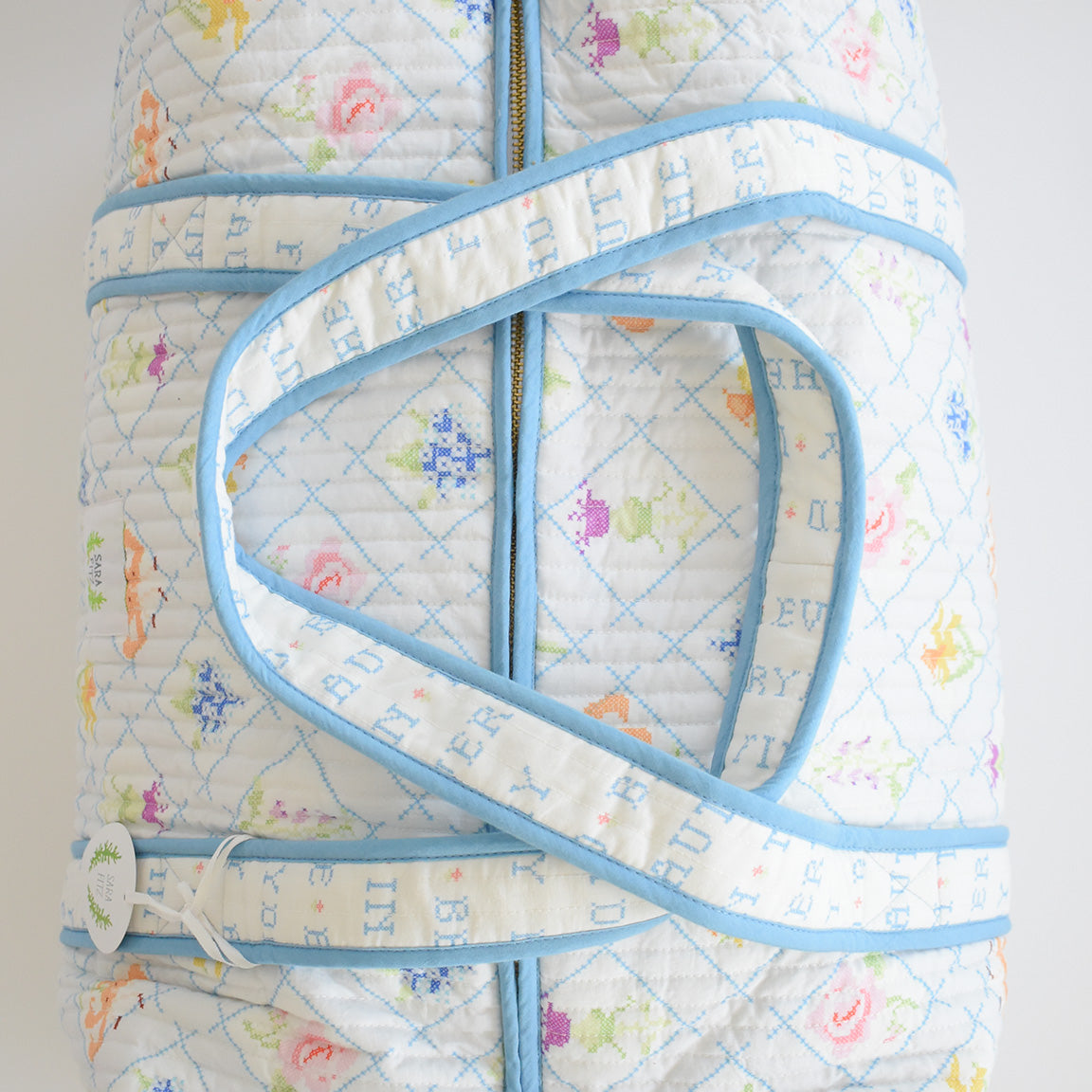 The Sara Fitz Weekender Tote, Cross Stitch Floral, is a cotton bag with blue trim, floral and cross-stitch designs, two blue-edged handles with blue lettering, and a white keychain—perfect as a stylish travel bag.