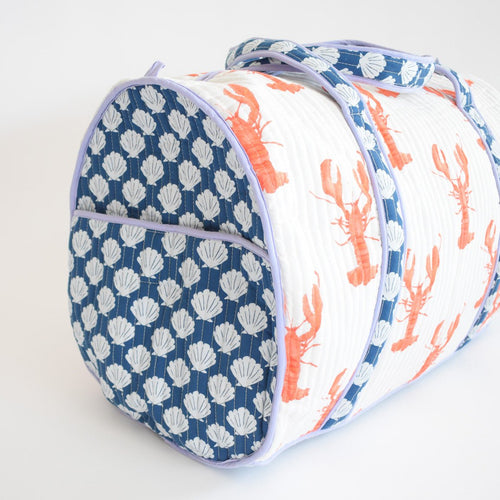 The Sara Fitz Weekender Tote, Lobster, is a spacious quilted cotton bag with blue handles, light purple piping, blue and white seashells on the end pocket, and fun orange lobsters on the white side panels.
