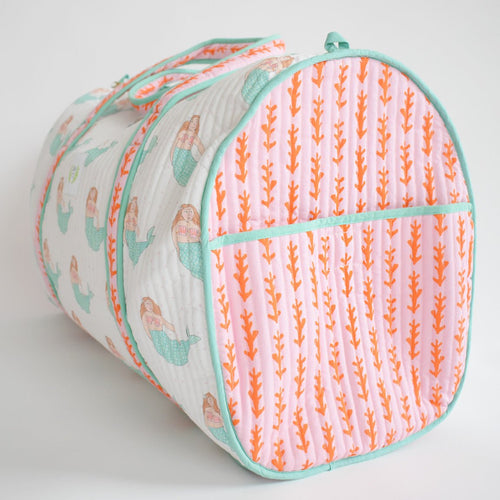 The Sara Fitz Weekender Tote, Mermaid, is a cylindrical quilted bag in light pink cotton with orange coral and mermaid designs, turquoise piping and handles, shown lying on its side against a white background.