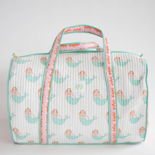 The Sara Fitz Weekender Tote, Mermaid, is a quilted cotton bag with turquoise handles and trim, featuring pastel mermaids and orange coral on the straps against a white background.