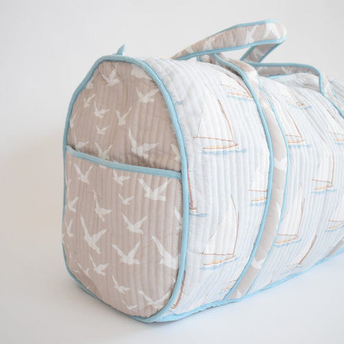 The Sara Fitz Weekender Tote, Sailboat, is a quilted travel bag with light blue trim and a pattern of white birds and sailboats on a light gray background, displayed against a plain white surface.