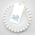 The Hydrangea Bloom Tags by Sara Fitz feature a white gift tag with yellow-white baker's twine and a watercolor light blue hydrangea with green leaves, displayed on a white scalloped plate.