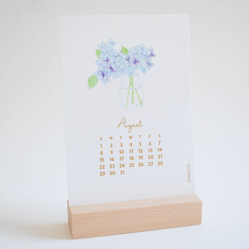 August page of a Sara Fitz 2027 Desk Calendar featuring a watercolor illustration of blue hydrangeas in a glass jar, set in a wooden stand.