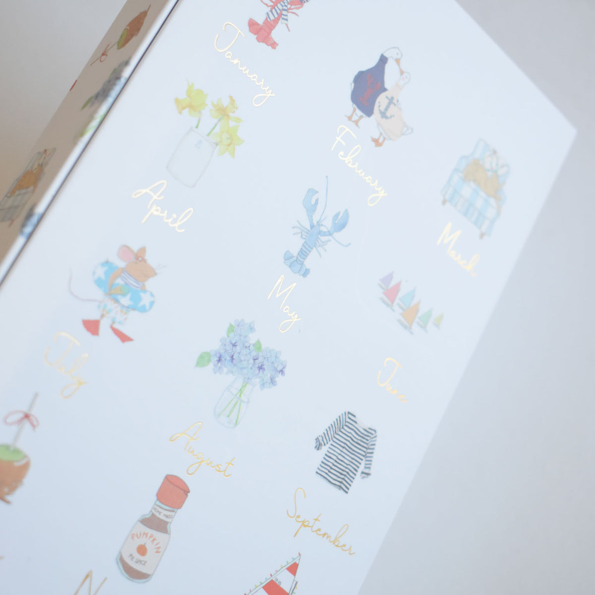 A 2027 Desk Calendar page featuring gold foil monthly labels and Sara Fitz illustrations of a lobster, geese, a mouse in a floatie, flowers, and a bottle of pumpkin spice.