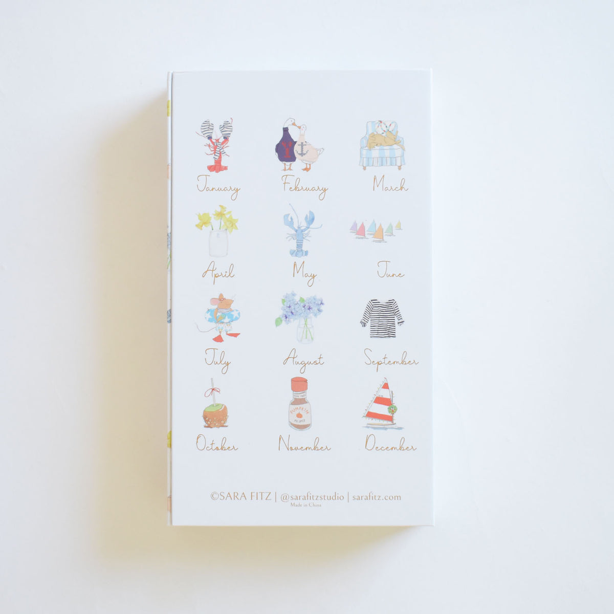 A rectangular 2027 Desk Calendar by Sara Fitz featuring twelve monthly pages, each illustrated with a seasonal watercolor motif.
