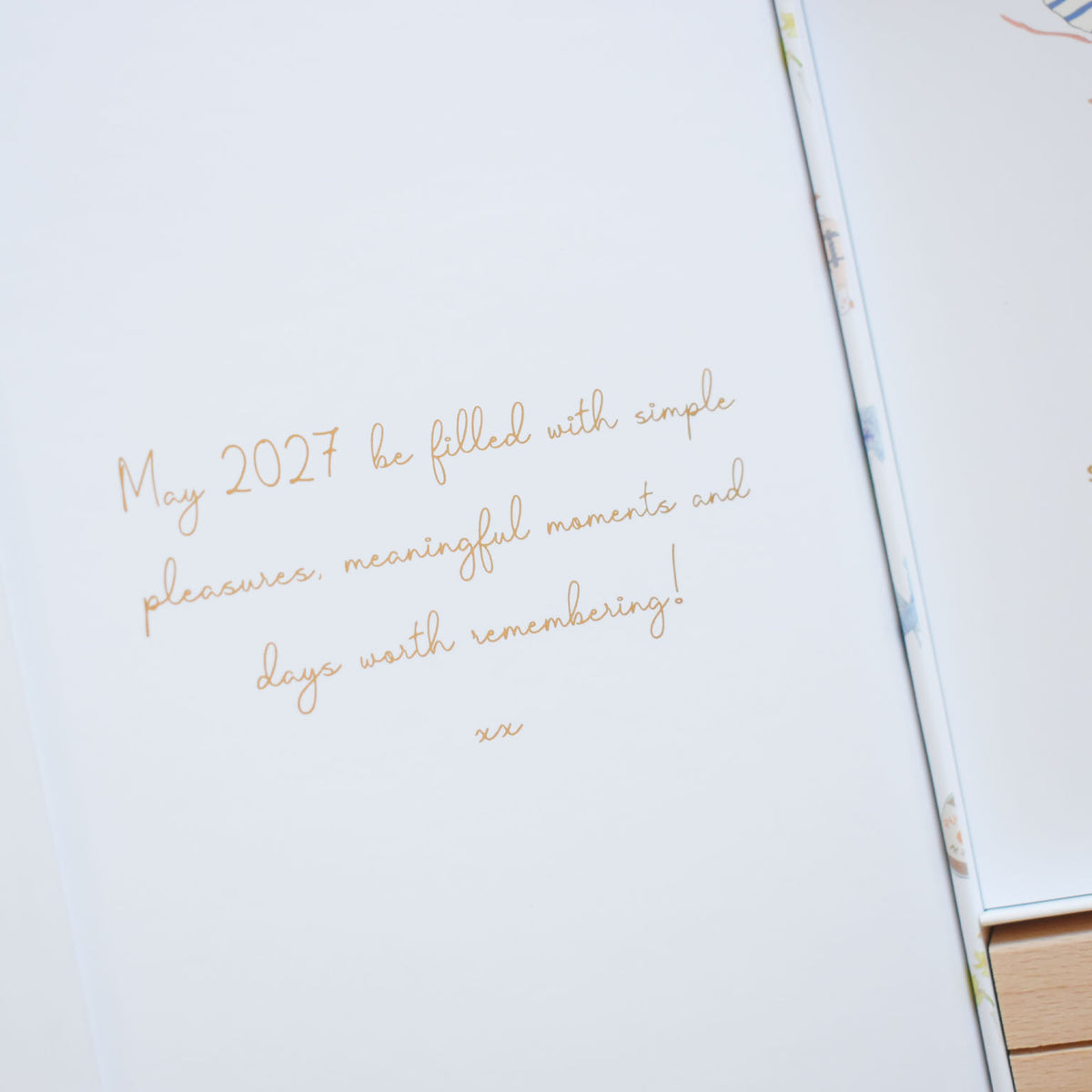 Close-up of a page from a Sara Fitz 2027 Desk Calendar featuring the handwritten-style inscription: May 2027 be filled with simple pleasures, meaningful moments and days worth remembering! xx.