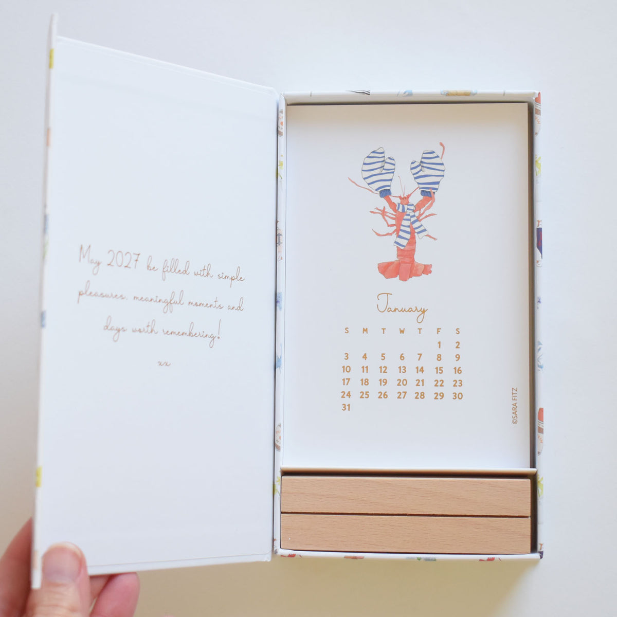 An open Sara Fitz 2027 Desk Calendar displaying a January page with a lobster wearing a scarf, resting in a wooden stand inside a keepsake box.