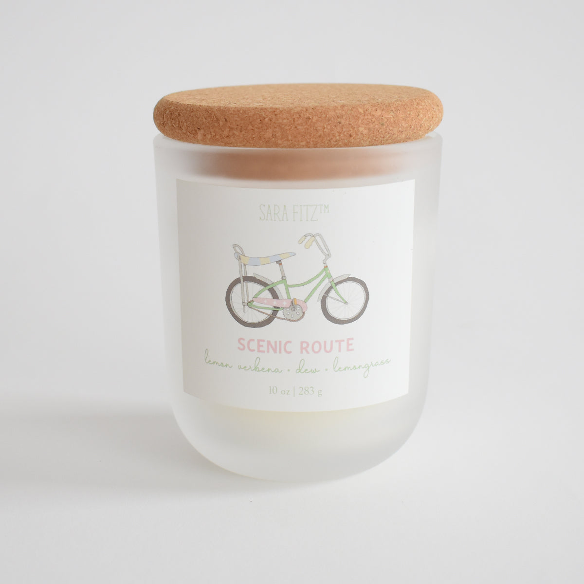The Sara Fitz Scenic Route Candle is hand-poured in Maine in a frosted glass jar with cork lid, featuring a bicycle label. Scented with lemon verbena, clove, and lemongrass; made from coconut soy wax, 10 oz (283 g).