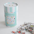 A light blue striped cylindrical container for the Seaside Puzzle, featuring an illustration of the seaside, resting next to a scattered pile of puzzle pieces.