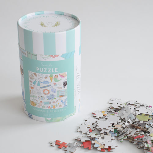 A light blue striped cylindrical container for the Seaside Puzzle, featuring an illustration of the seaside, resting next to a scattered pile of puzzle pieces.