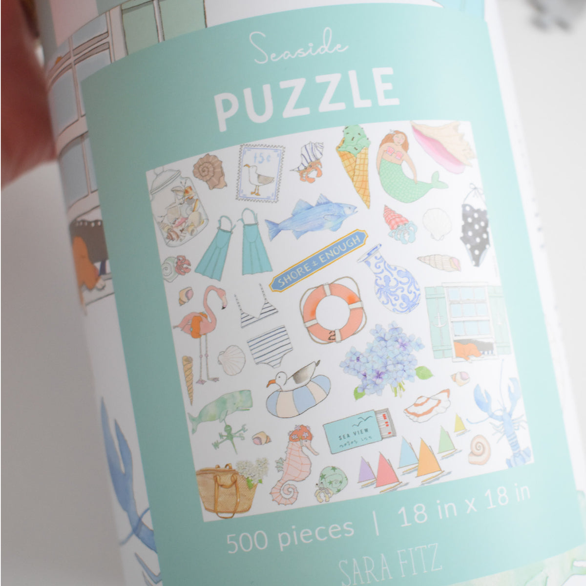 A cylindrical container for a 500-piece Seaside Puzzle featuring colorful illustrations of seashells, sea life, and ice cream with text reading Seaside PUZZLE, 18 in x 18 in, in a vibrant beach collage.