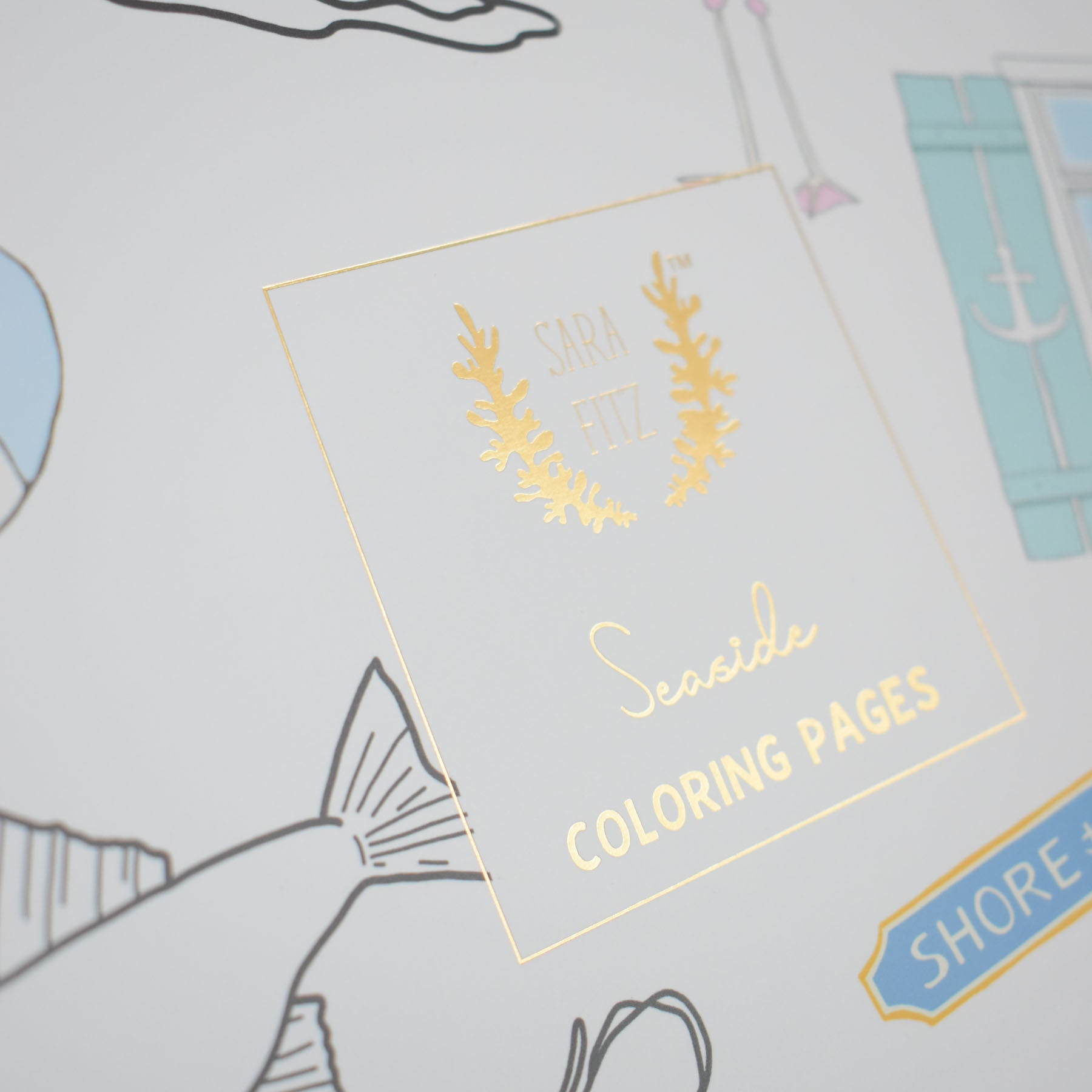 Close-up of the Coloring Book Seaside cover by Sara Fitz, featuring gold foil text and partially visible illustrations of fish, a window, and nautical items inspired by New England.