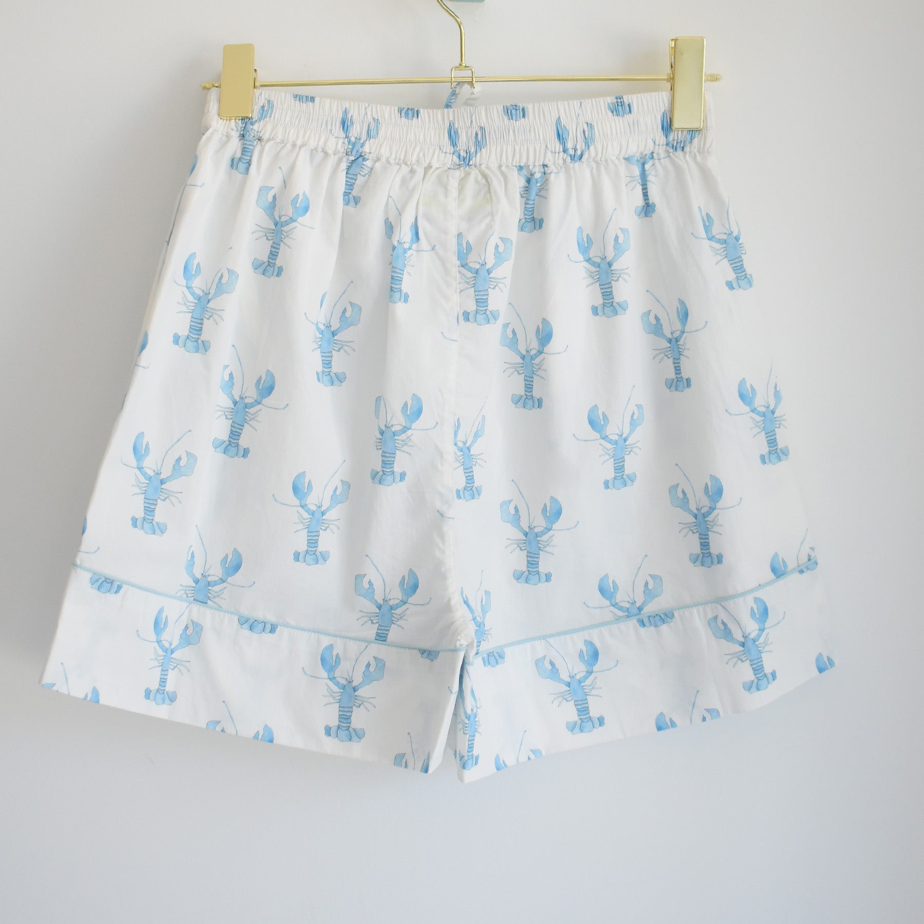 White shorts with an elastic waistband, adorned with a light blue lobster pattern, hang on a gold hanger against a plain white background—cozy and stylish, these bottoms are part of the Sara Fitz Blue Lobster Short Pajama Set, perfect for bringing coastal pajama style to your nightly routine.