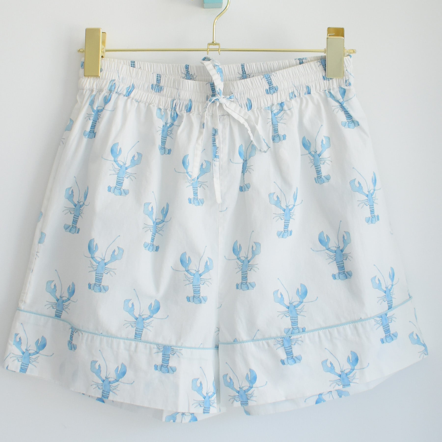 White shorts featuring a light blue lobster pattern, elastic waistband, and drawstring, displayed on a gold hanger against a plain white background—these stylish bottoms are part of the Sara Fitz Blue Lobster Short Pajama Set, ideal for adding coastal charm to your relaxation routine.