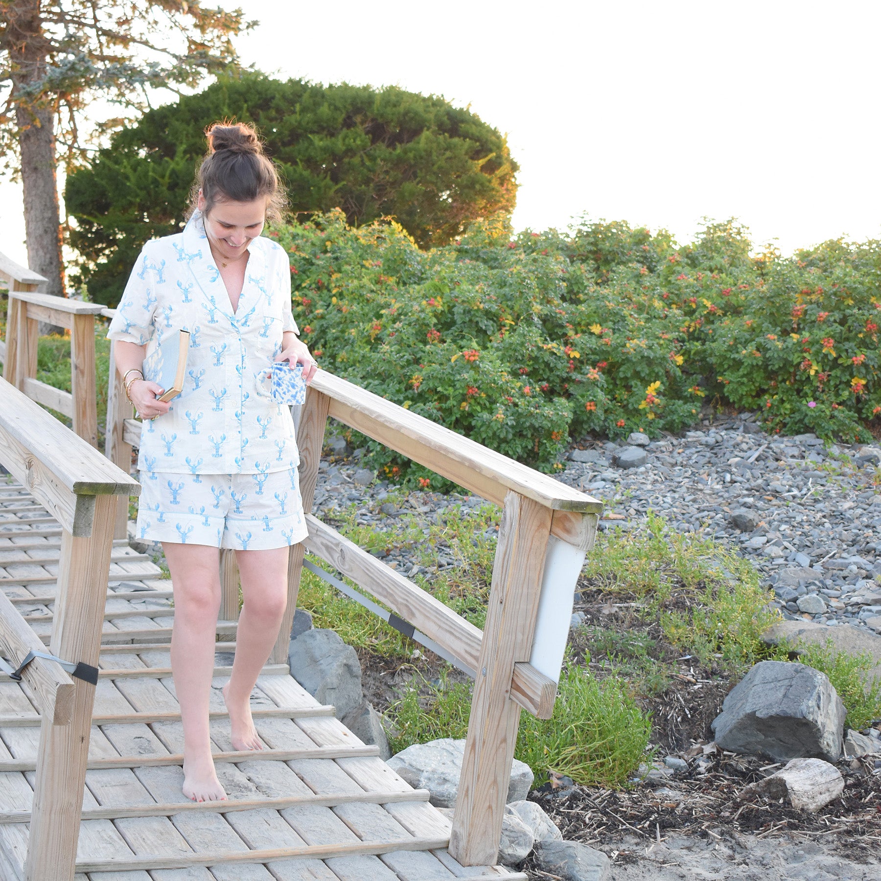 A woman in the Sara Fitz Blue Lobster Short Pajama Set walks barefoot down a wooden ramp toward a rocky area, holding a book and a cup, with greenery and flowers in the background.