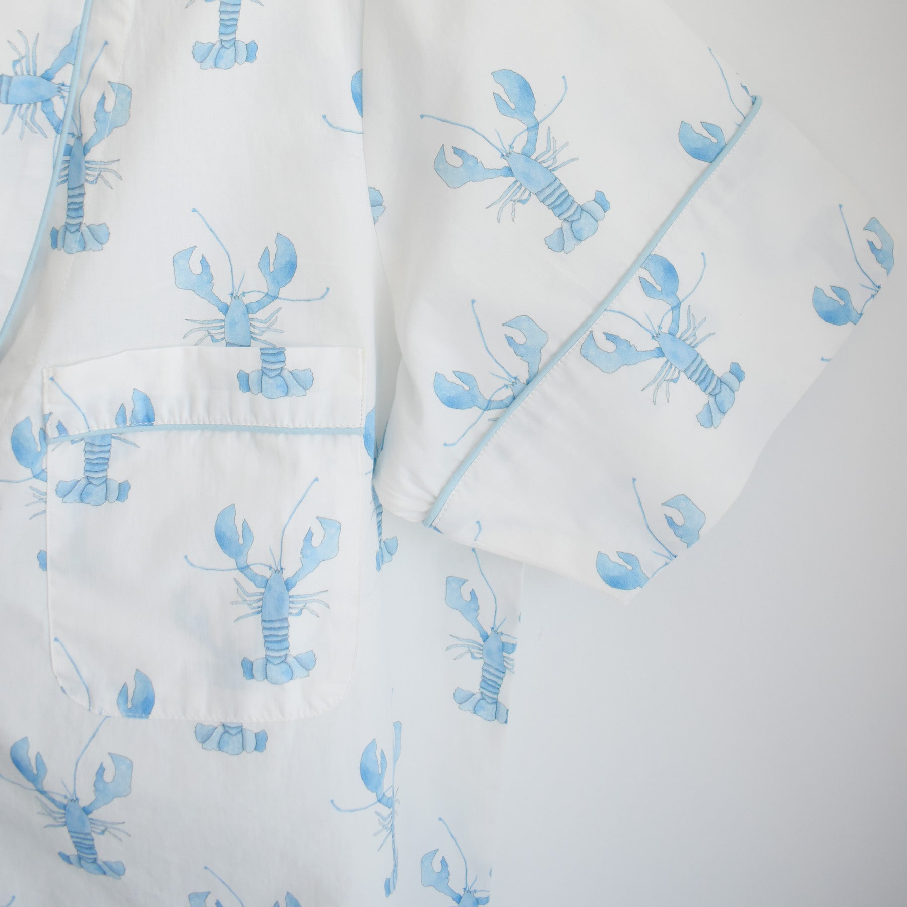 The Blue Lobster Short Pajama Set from Sara Fitz features a white shirt adorned with a charming blue lobster print, a front pocket, and blue piping along the pocket and sleeve edges. Crafted from soft cotton, this coastal-inspired pajama set combines comfort and playful style for cozy nights in.