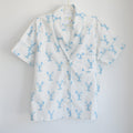 The Blue Lobster Short Pajama Set by Sara Fitz features a short-sleeved white button-up shirt adorned with a playful blue lobster print, a notched collar, and a front pocket on the left chest. Displayed against a plain white background, this set is perfect for those seeking coastal-inspired pajamas with whimsical charm.