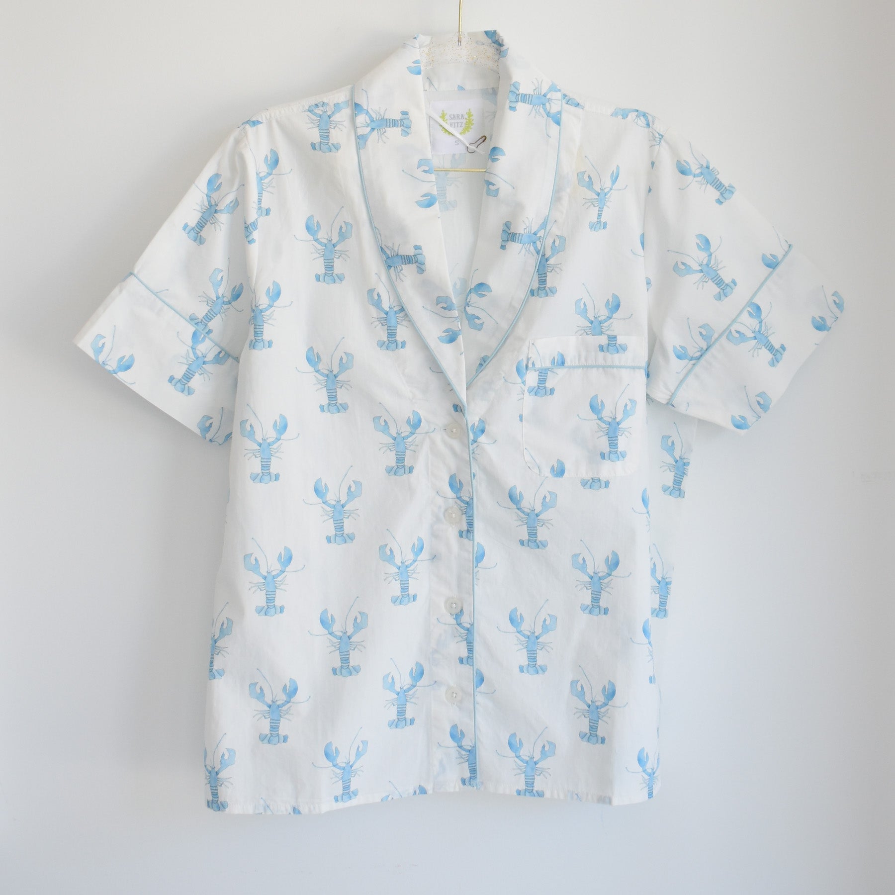 The Blue Lobster Short Pajama Set by Sara Fitz features a short-sleeved white button-up shirt adorned with a playful blue lobster print, a notched collar, and a front pocket on the left chest. Displayed against a plain white background, this set is perfect for those seeking coastal-inspired pajamas with whimsical charm.