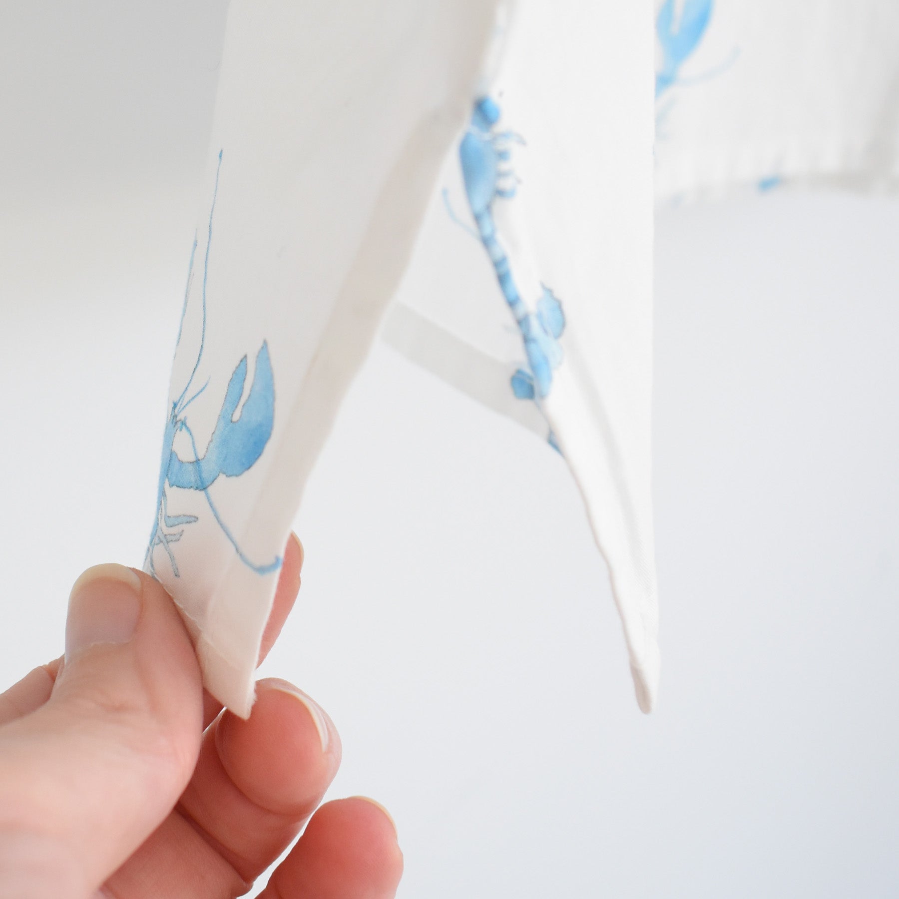 A hand holds the corner of the Sara Fitz Blue Lobster Sleep Shirt, featuring a white cotton fabric adorned with a light blue lobster print, against a plain, light background—a cozy addition to any women’s sleepwear collection.