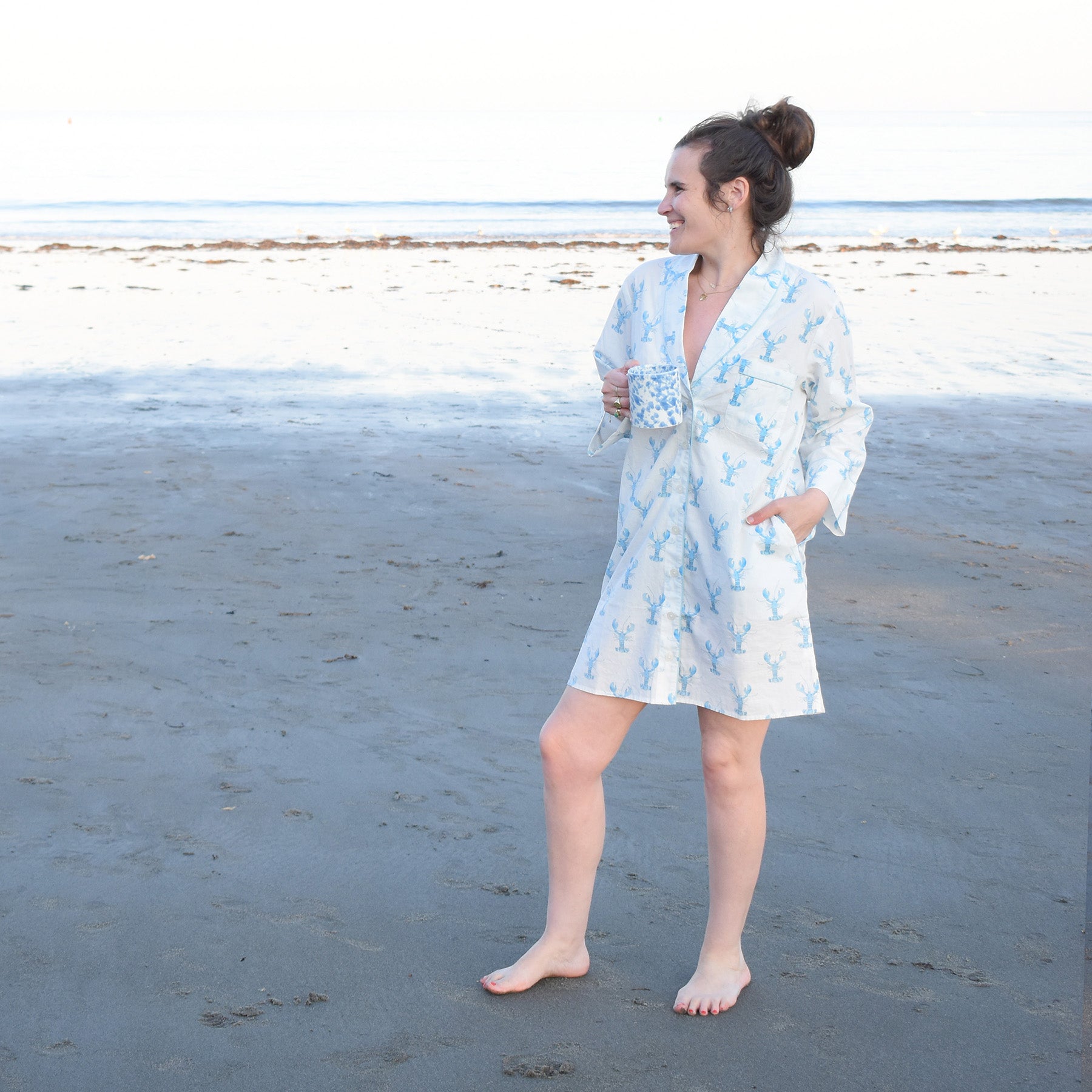 A woman in a patterned robe stands barefoot on a sandy beach, smiling and holding a mug, with calm ocean water in the background. Beneath her robe, she wears the Sara Fitz Blue Lobster Sleep Shirt—an essential piece of women’s sleepwear perfect for relaxed mornings by the sea.