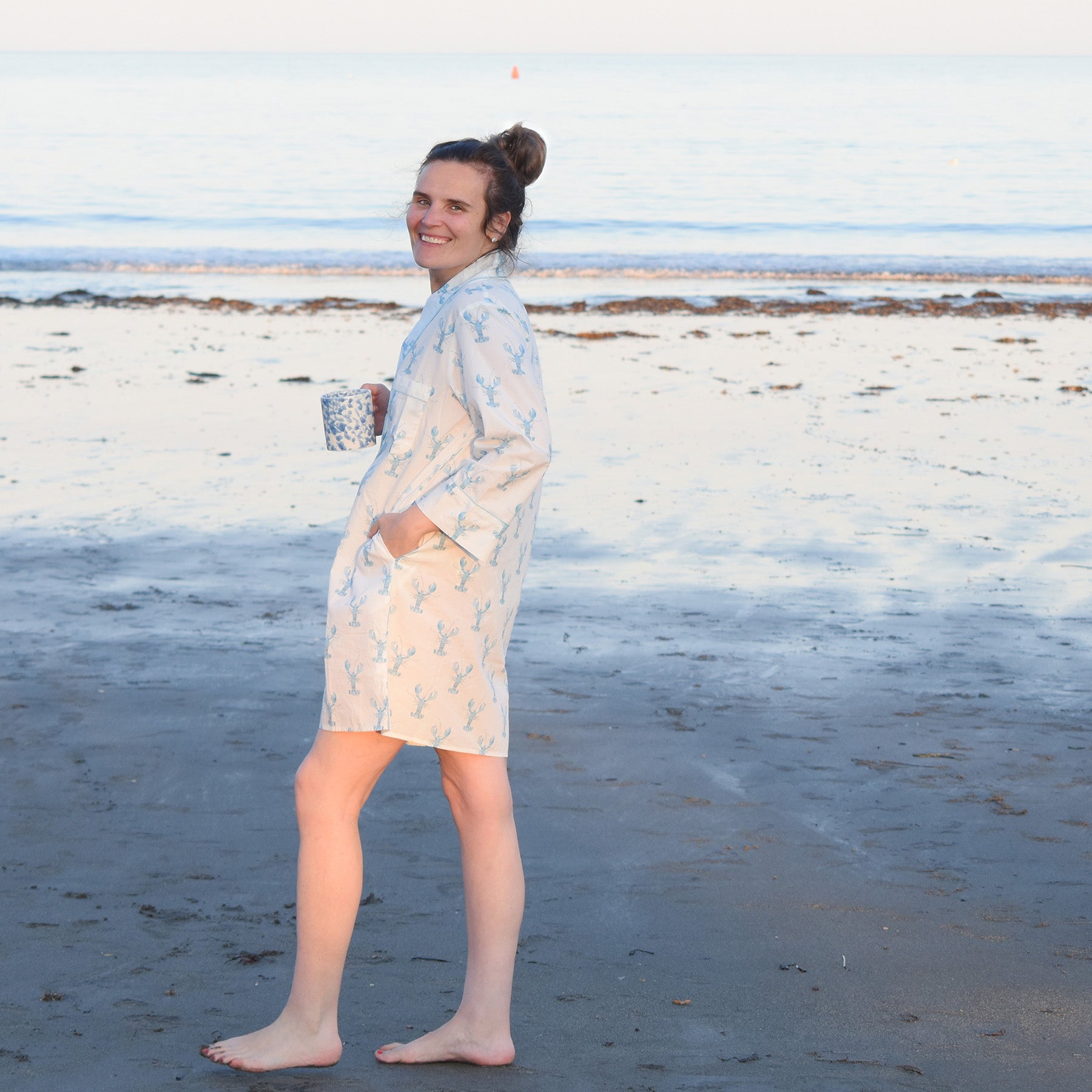 A person stands barefoot on a sandy beach, smiling at the camera while holding a mug and wearing a patterned light robe over the Sara Fitz Blue Lobster Sleep Shirt. The sea and sky create a calm backdrop.