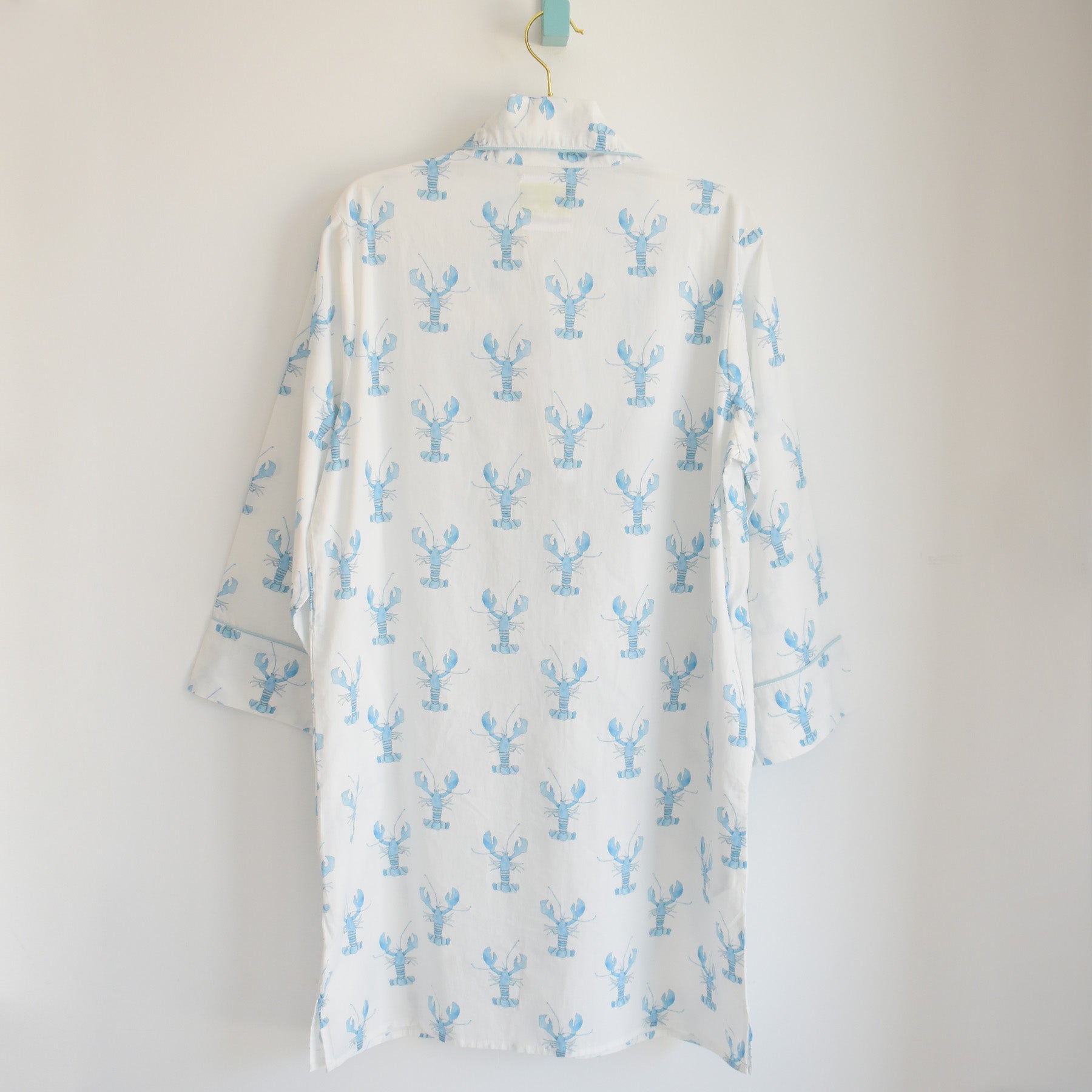 A “Blue Lobster Sleep Shirt” by Sara Fitz, featuring a light blue lobster pattern on white fabric, is hanging on a hook against a plain white wall with its back displayed to the viewer.